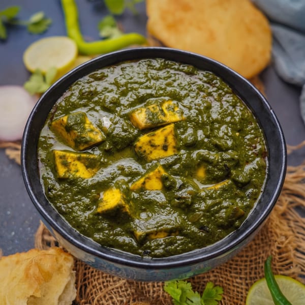 Bowl of Indian palak paneer with golden paneer cubes in creamy spinach sauce, garnished with lime, onion, chili, and cilantro.