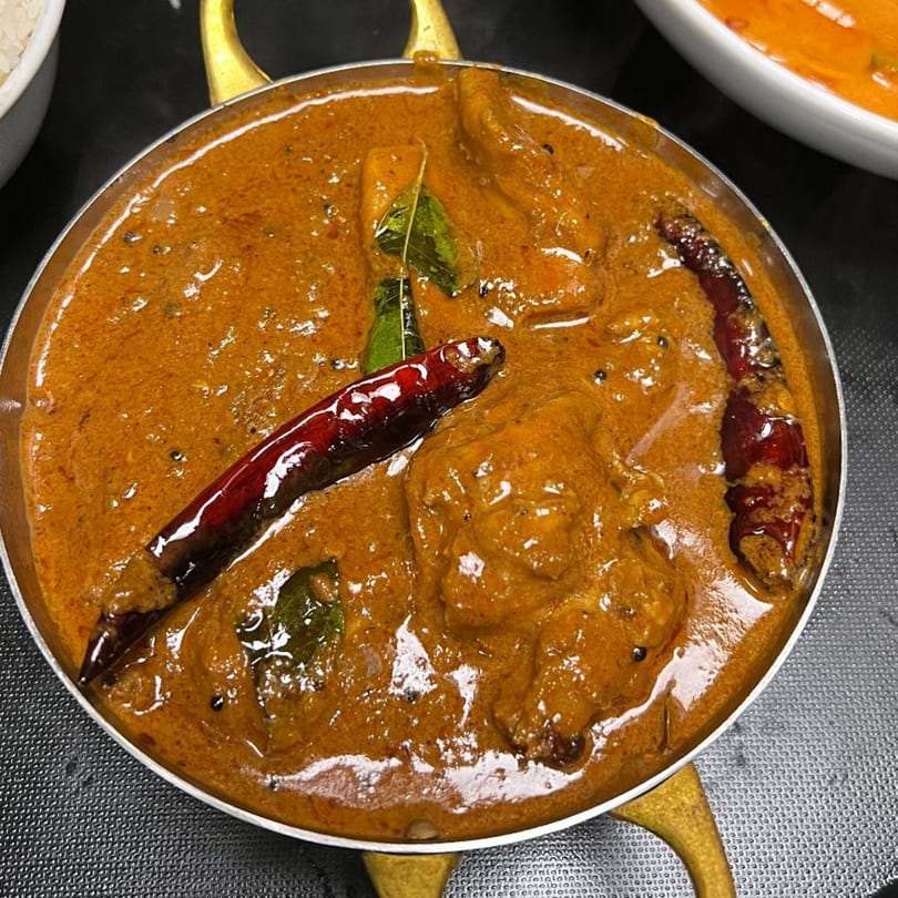 A close-up of a rich, orange Indian curry with whole red chilies and green leaves in a metal bowl.