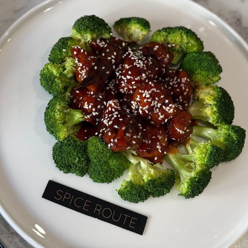 Indian-style chicken with a dark glaze and sesame seeds, surrounded by broccoli florets on a white plate.