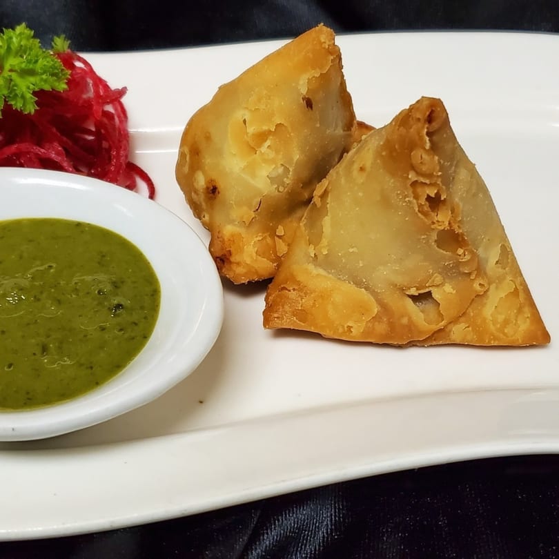 Two golden-brown Indian samosas with green chutney and red pickled onions on a white plate.